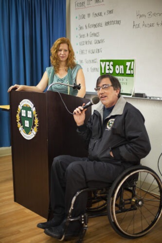 Dale Sky Jones stands at a podium with Richard Lee speaking into a microphone from his wheelchair in front.