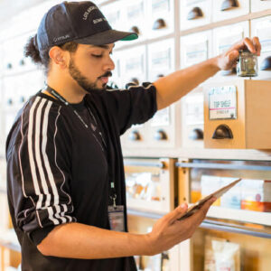 Male budtender with tablet in one hand inspecting inventory. In other hand, a glass jar of cannabis flower.